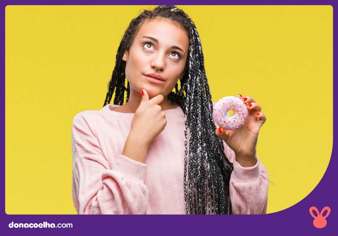 Pessoa com a mão no queixo em formato de dúvida, enquanto segura uma rosquinha com a outra mão